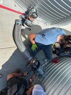 Two people crouch by a motorcycle wheel in a garage, with tools and a large black hose nearby.