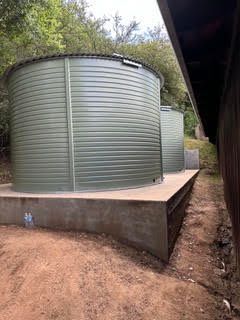 Two large green water tanks on a concrete platform beside a dirt path and building wall.