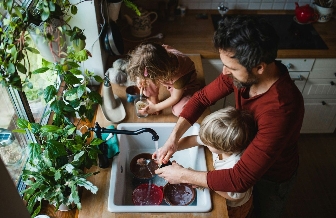 Two people and a child washing dishes together at a kitchen sink with a red basin.