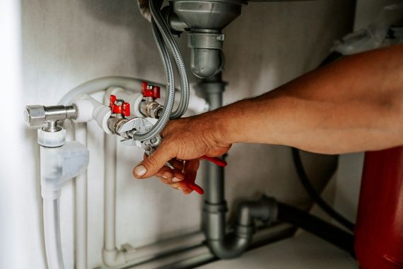 Hand turning a valve under a sink with exposed plumbing and red shutoff handles