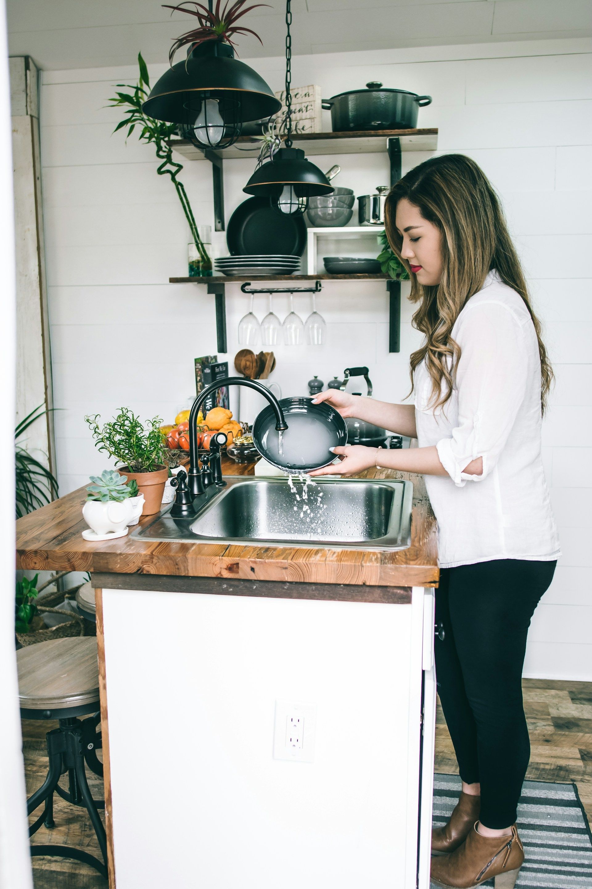 Woman washing dishes at a kitchen sink with shelves and plants in the background