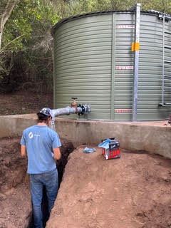 Man in blue shirt inspecting pipes beside a large green water tank outdoors
