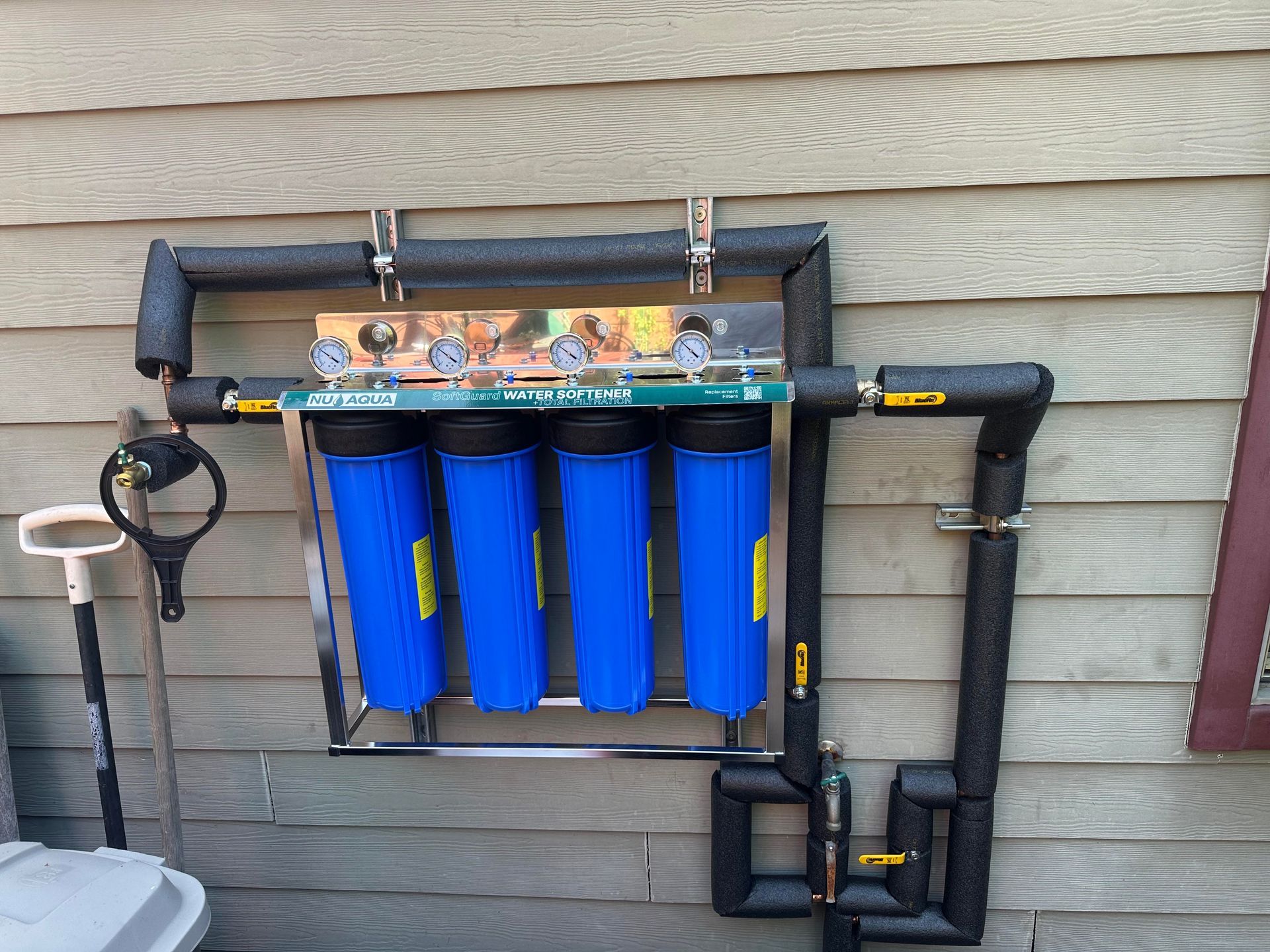 Wall-mounted blue water filtration system with pipes and gauges outside a house.