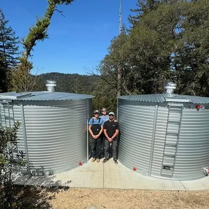 Two people standing between two large silver water tanks outdoors, with trees and hills in the background.