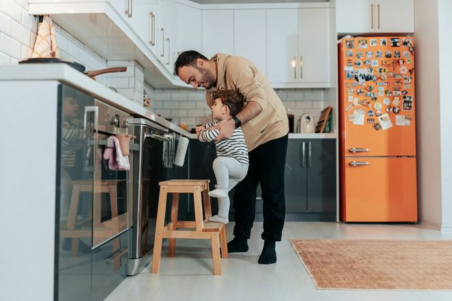Adult helping a child stand at a kitchen counter near an orange fridge