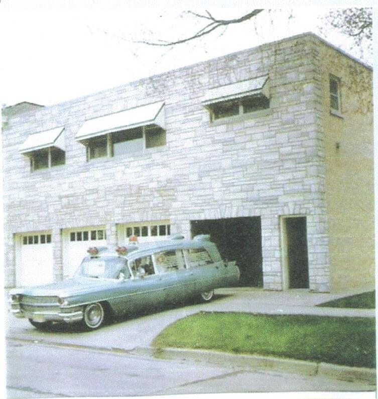 Light blue hearse parked in front of a stone building with three garages and awnings.