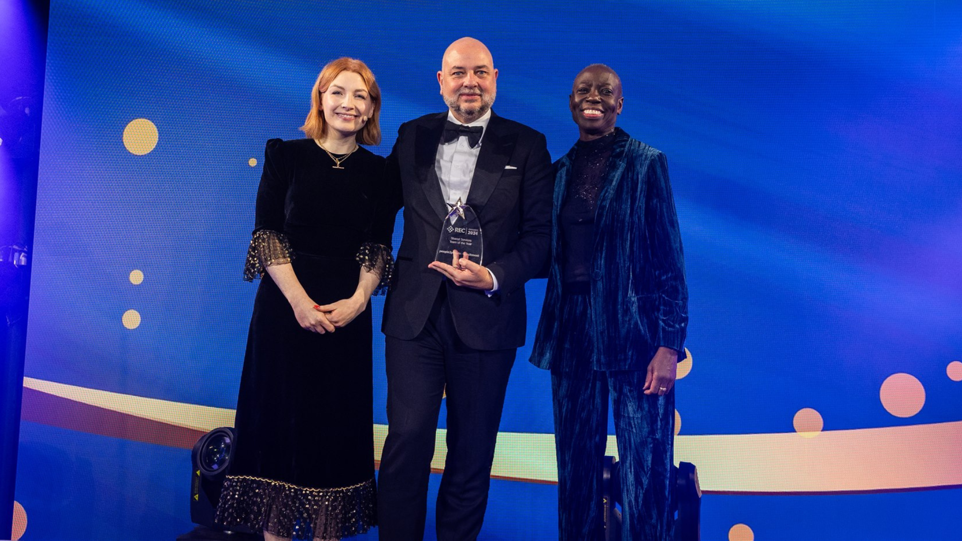 Three people stand on a stage with a blue background, one holding an award.