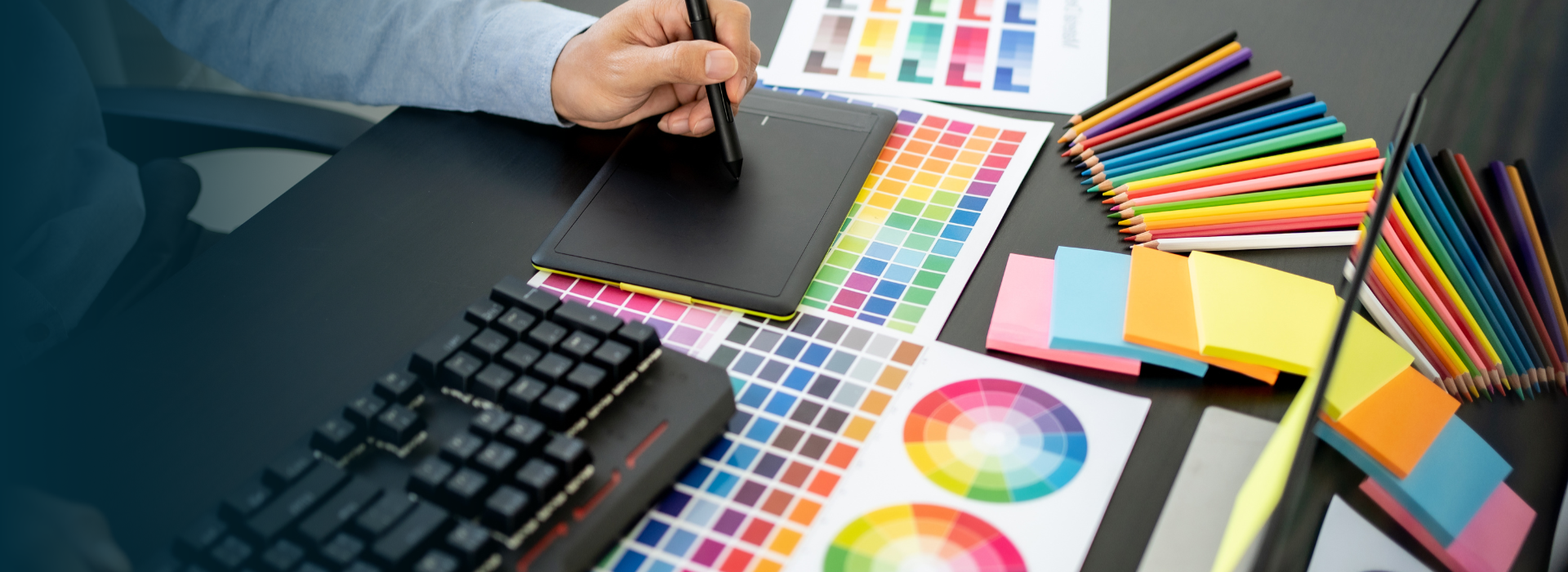 A graphic designer at a desk uses a stylus on a drawing tablet, surrounded by color swatches and samples.