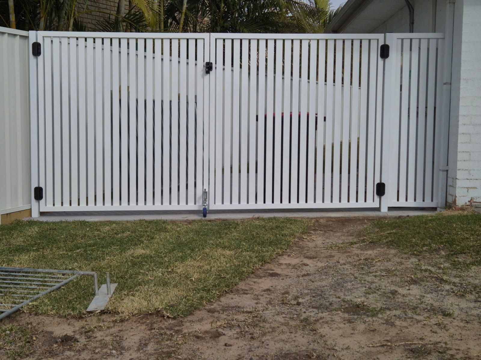 White vertical-slat driveway gate in front of a house with grass and dirt path foreground — FBM Fabrication In Dapto, NSW