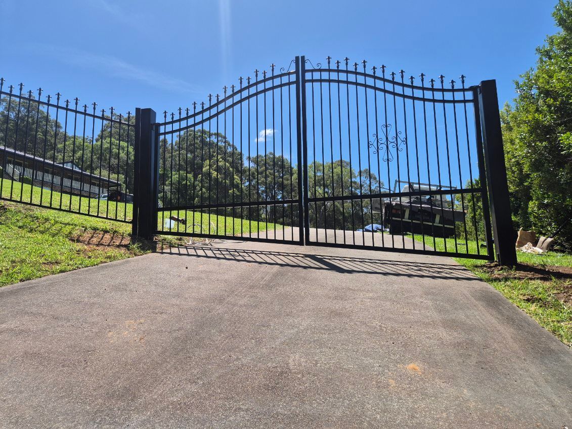 Black wrought-iron gate at the end of a driveway under a bright blue sky — FBM Fabrication In Dapto, NSW