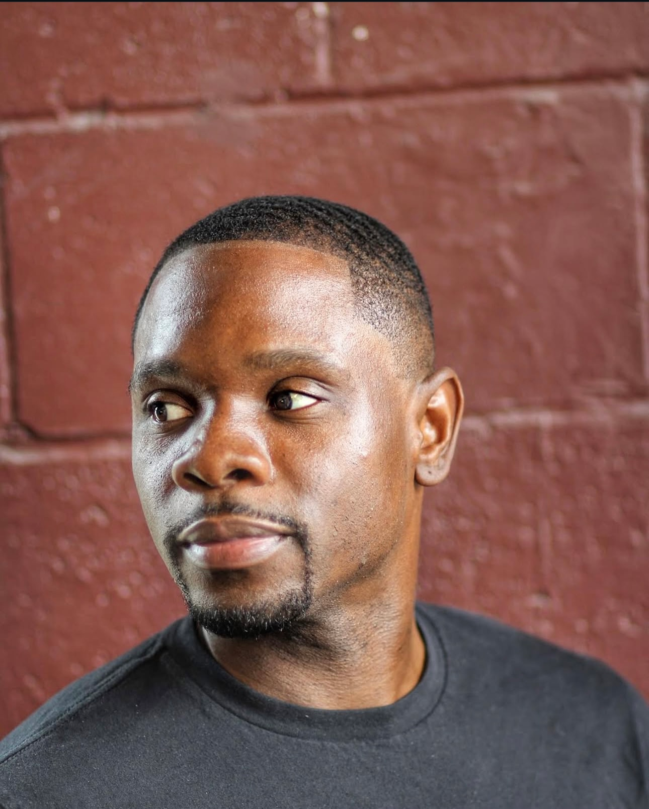 Man with a fade haircut and beard, against a brick wall.