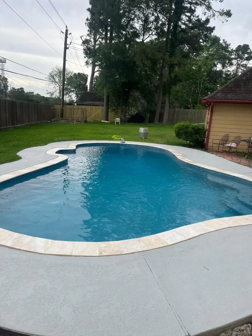 An outdoor swimming pool with blue water and light-colored stone coping, surrounded by grey decking and a grassy yard.