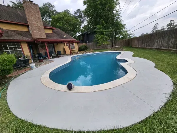 A backyard featuring an irregularly shaped blue swimming pool surrounded by a grey concrete deck near a house and lawn.