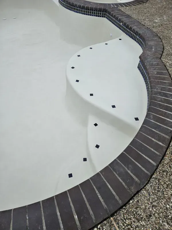 An empty swimming pool with white plaster walls, rounded steps with dark accent tiles, and a dark brick coping edge.
