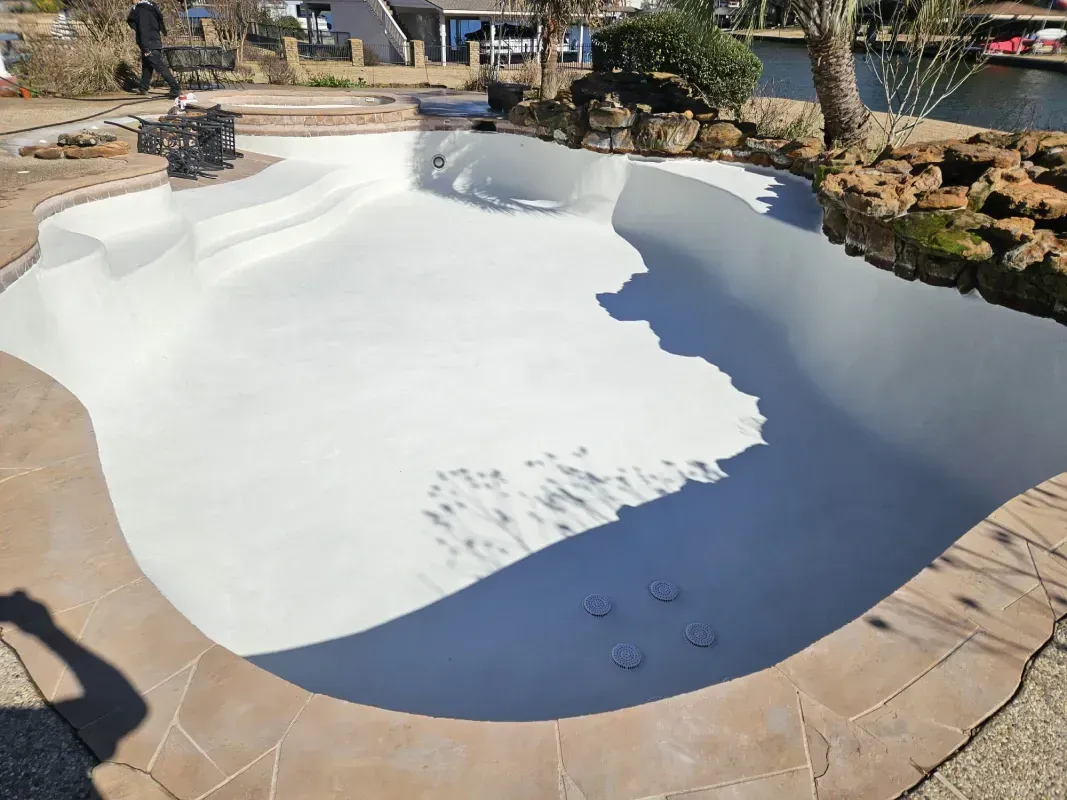 An empty, freshly resurfaced white swimming pool with stone coping, surrounded by a yard and a view of a canal.