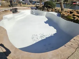 An empty, freshly resurfaced white swimming pool with stone coping, surrounded by a yard and a view of a canal.