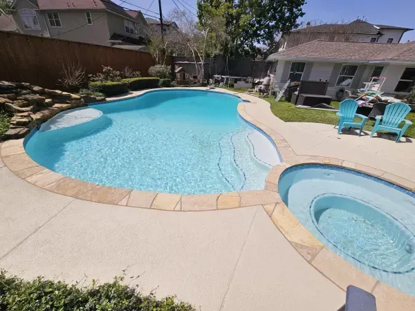 A backyard swimming pool with a stone border, an attached circular spa, and two light blue chairs on the concrete patio.