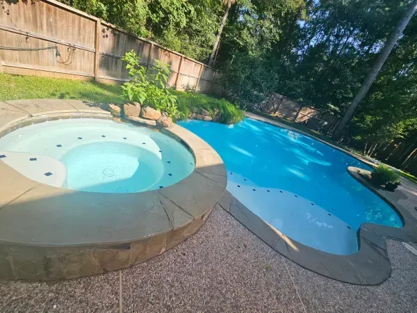 A backyard swimming pool and a round stone hot tub, both filled with clear blue water, set on a textured patio.