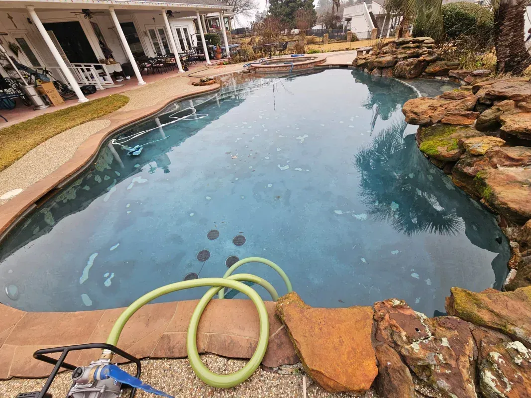 A backyard swimming pool with a rock feature and a pump with a green hose in the foreground.