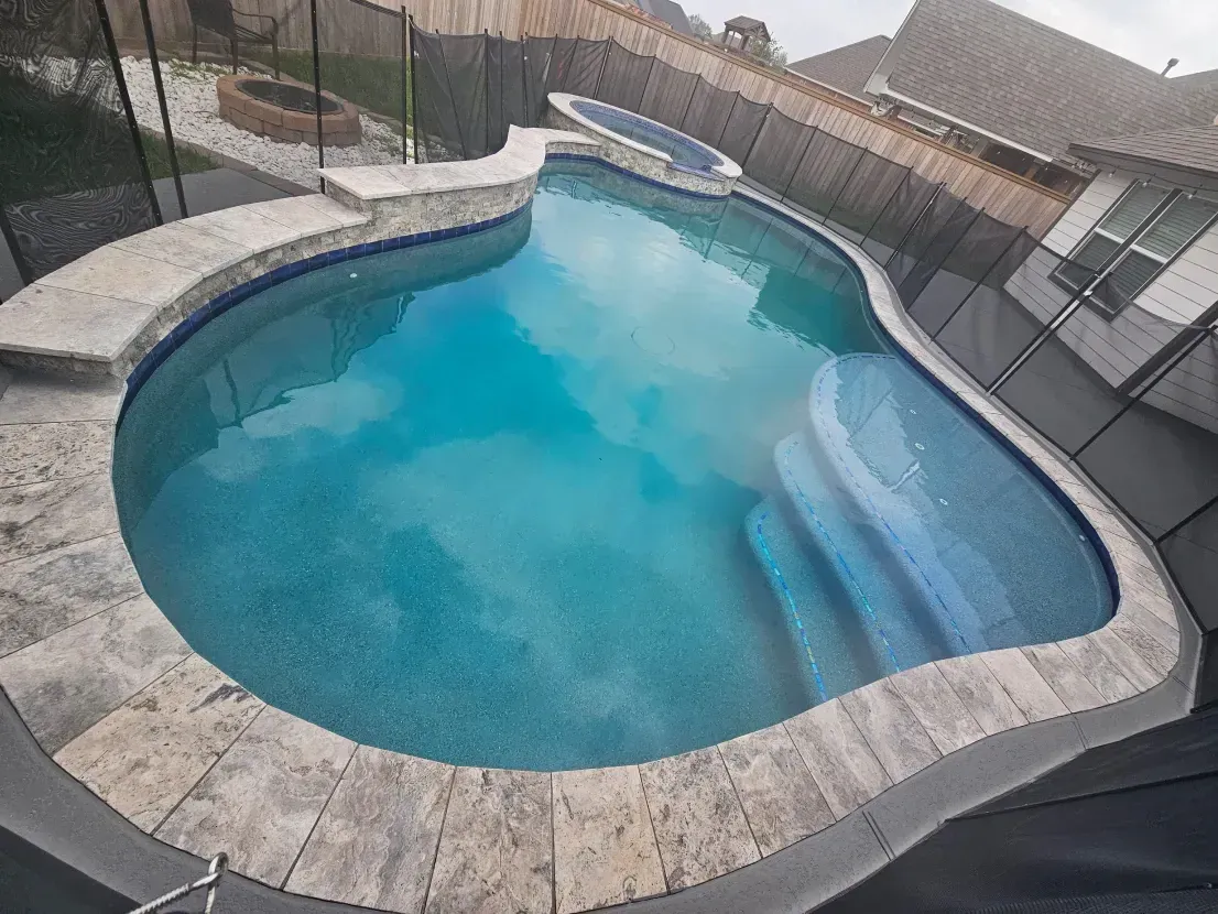 A curved backyard swimming pool with light blue water, stone coping, and built-in steps, surrounded by a safety fence.