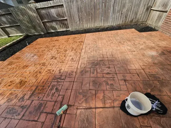 A stamped concrete patio is being refinished, showing a contrast between the darker, treated area and the lighter, dry side.