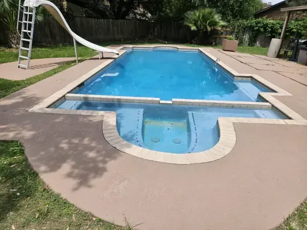 A rectangular swimming pool with an attached shallow lounging area and a white slide on a concrete patio in a yard.