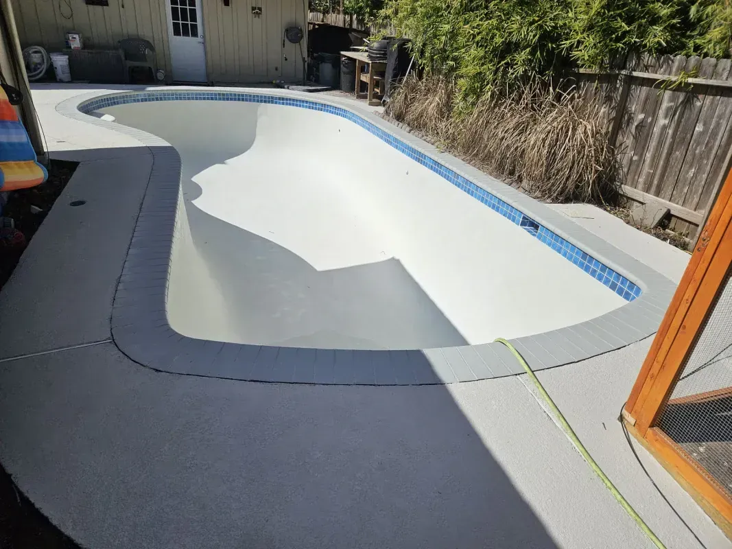 An empty, light-colored backyard swimming pool with blue tile trim, surrounded by a grey concrete deck.