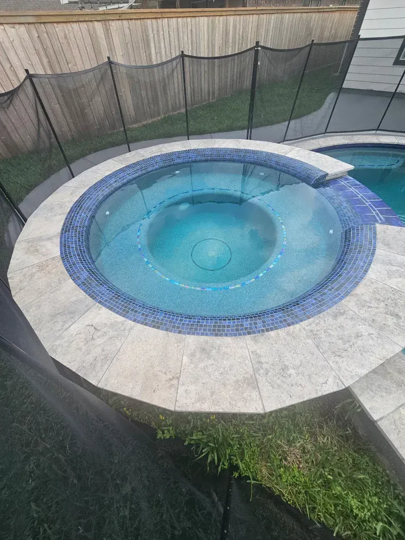 A round hot tub with blue tile edges and light-colored stone pavers, positioned next to a pool behind a black safety fence.
