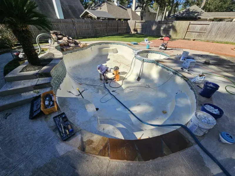 An empty backyard swimming pool undergoing resurfacing, surrounded by tools, buckets, and construction equipment.
