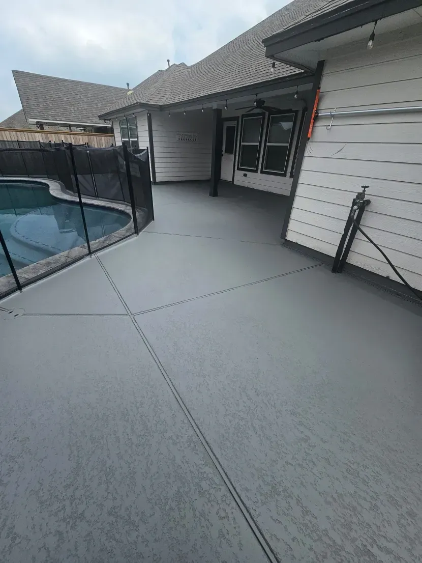 A gray-coated concrete patio next to a swimming pool with a safety fence outside a residential house.