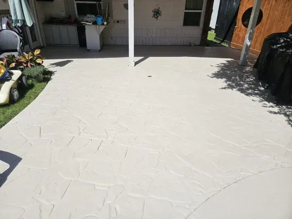 A light-colored patio with a stone-textured pattern, partially covered by a roof and surrounded by a yard.