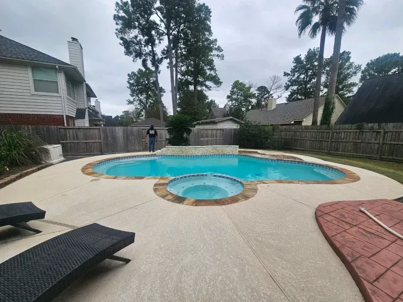 Backyard swimming pool with an attached spa, light-colored concrete decking, and a person standing near the wooden fence.