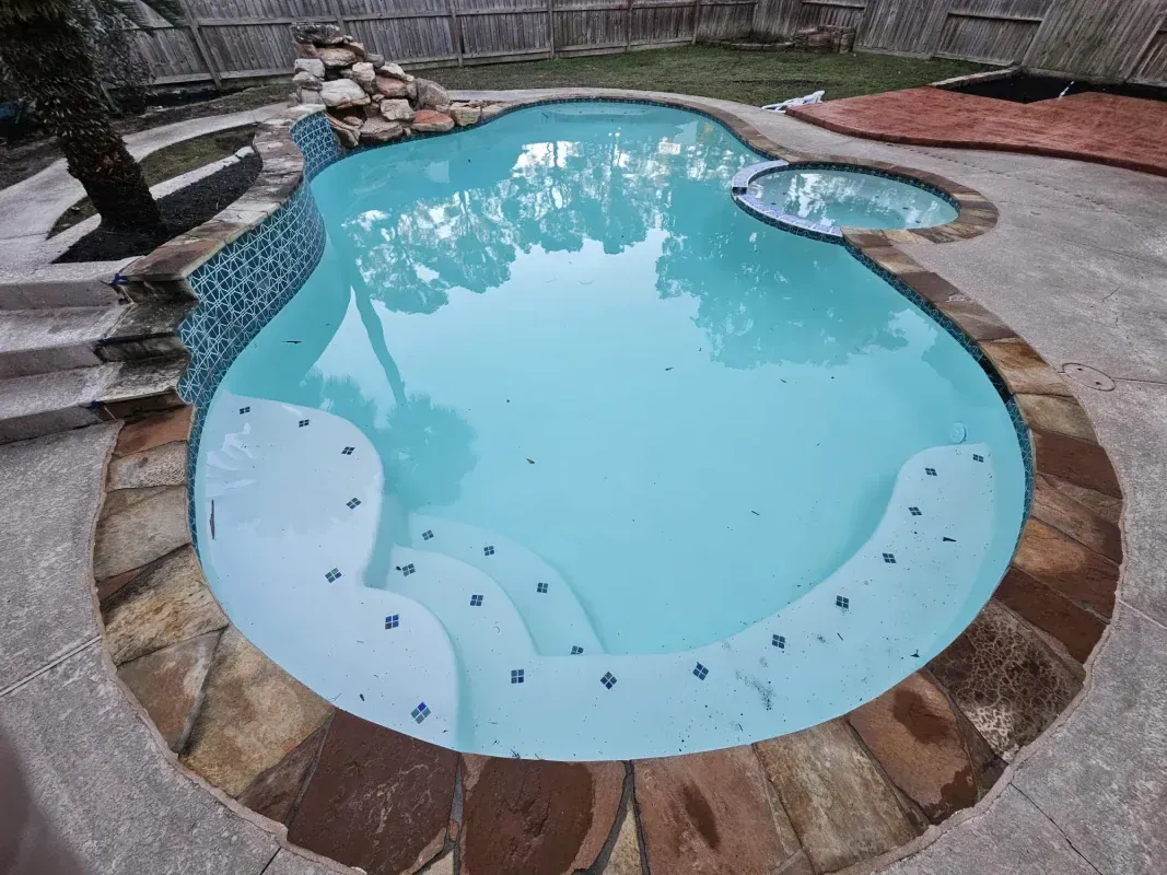 A bright blue swimming pool with a stone border, an attached hot tub, and stairs leading into the water.
