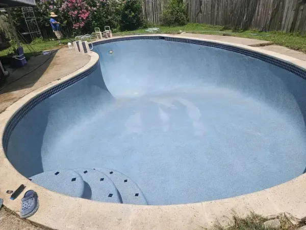 An empty, blue, kidney-shaped swimming pool with tiered steps, viewed from a patio on a sunny day.