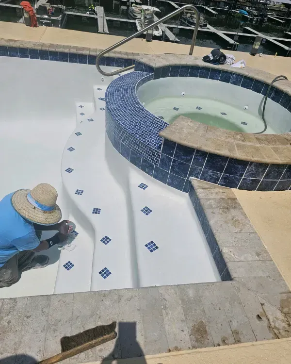 A person wearing a straw hat works on the tiled steps of an empty swimming pool next to a circular spa.