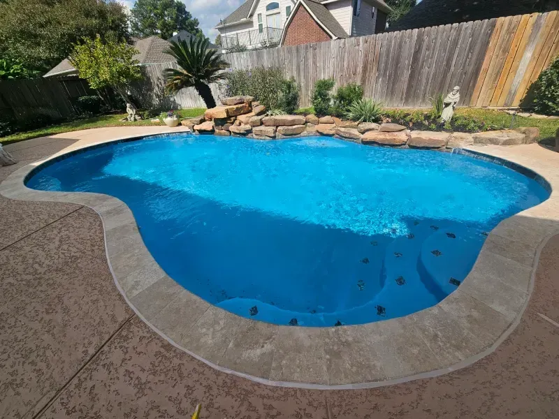 A free-form swimming pool with a rock water feature, surrounded by a stone patio in a sunny backyard.