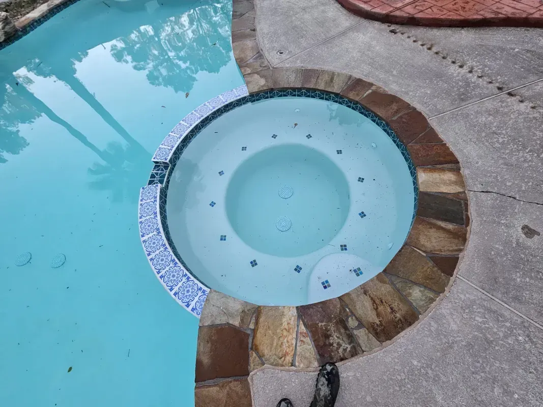 A round in-ground spa adjacent to a swimming pool, both featuring light blue water and stone and tile coping.