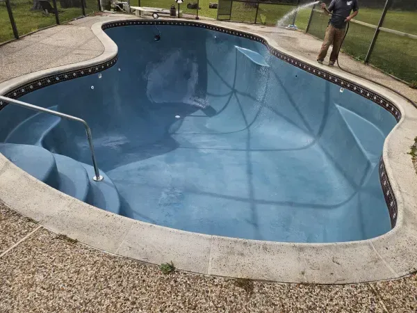 An empty, light blue, kidney-shaped swimming pool with built-in stairs being filled by a person with a hose.