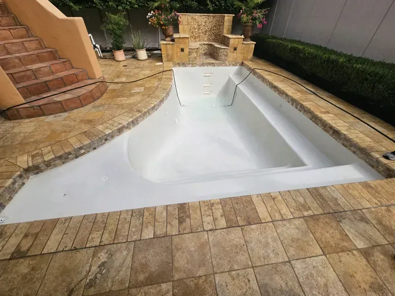 A white, empty fiberglass pool set into a stone patio with steps to the left and a stone bench feature in the background.