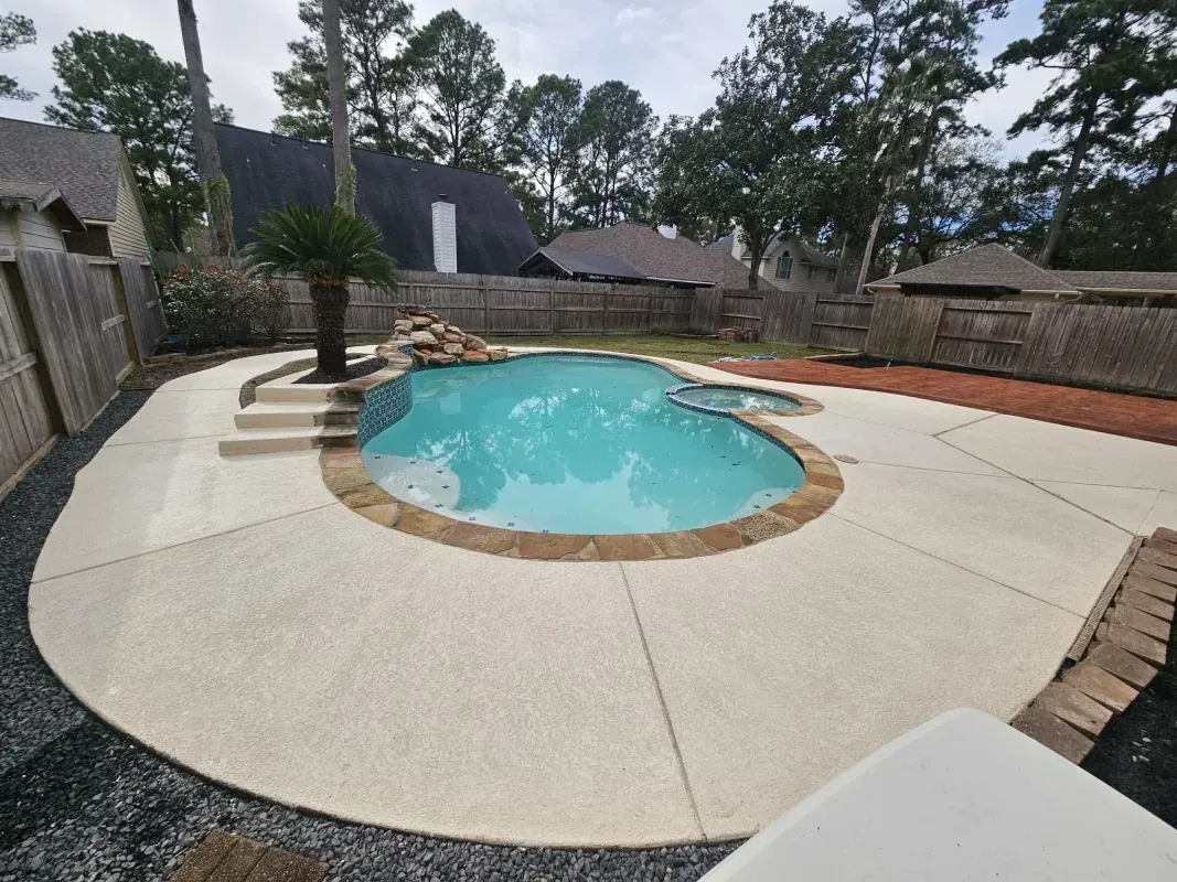 A backyard swimming pool with a small spa, palm tree, rock feature, and light-colored concrete deck, enclosed by a fence.