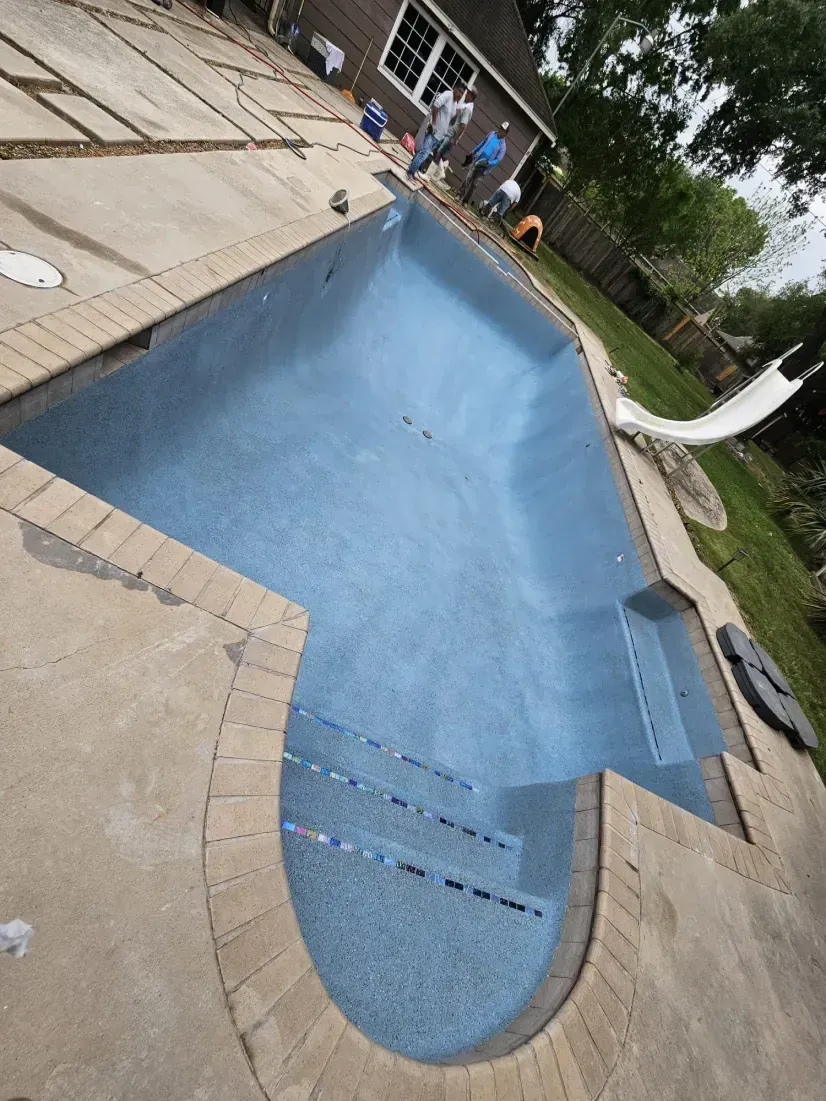An empty, freshly resurfaced blue swimming pool with stairs, surrounded by a stone patio and a backyard with a slide.