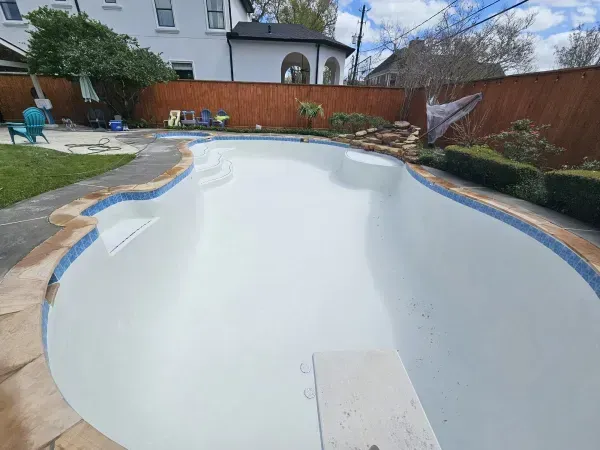Empty white backyard swimming pool with tiled edges, stone coping, and a diving board on a sunny day.