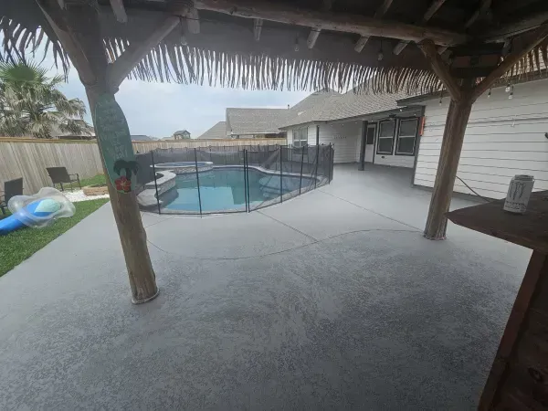 A grey patio with a swimming pool enclosed by a black mesh safety fence under a thatched-roof structure.