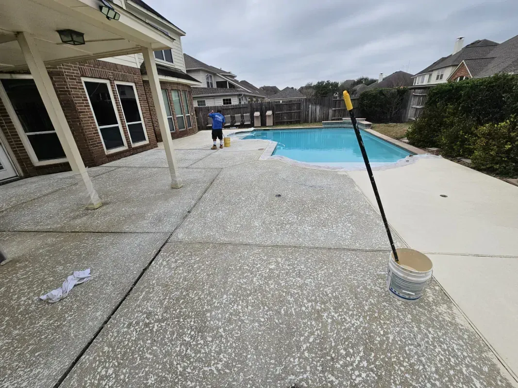A person stands on a patio near a pool, holding a long-handled brush next to a bucket on a residential backyard deck.