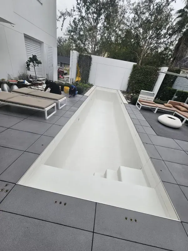 A narrow, empty rectangular swimming pool with steps, surrounded by gray patio tiles and outdoor lounge furniture.