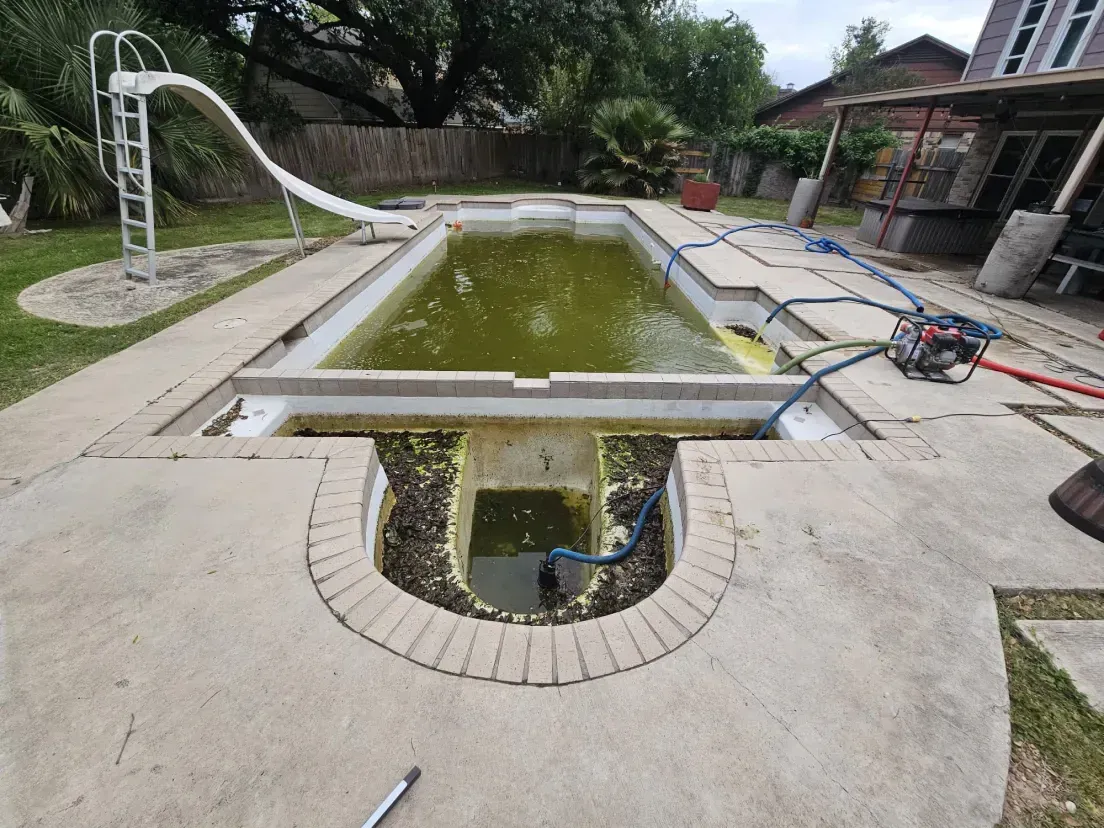 An outdoor backyard swimming pool with murky green water, a white slide, and a filtration system visible on the deck.