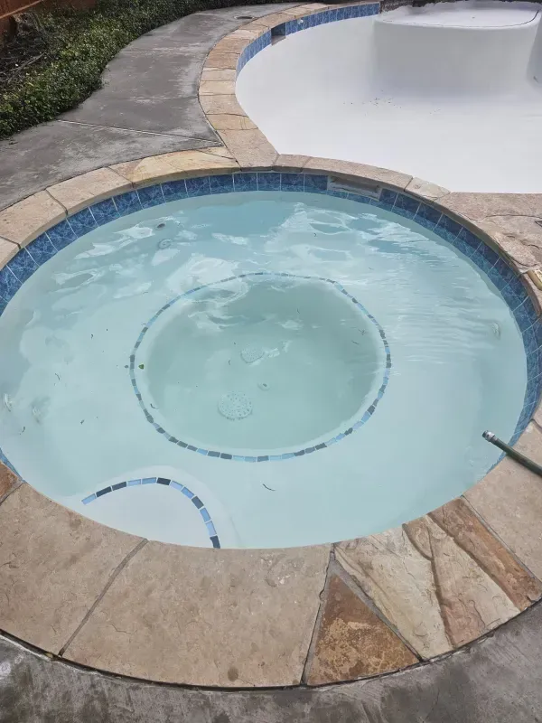 A round, light-colored hot tub with blue mosaic tile trim, situated next to an adjacent, larger swimming pool.