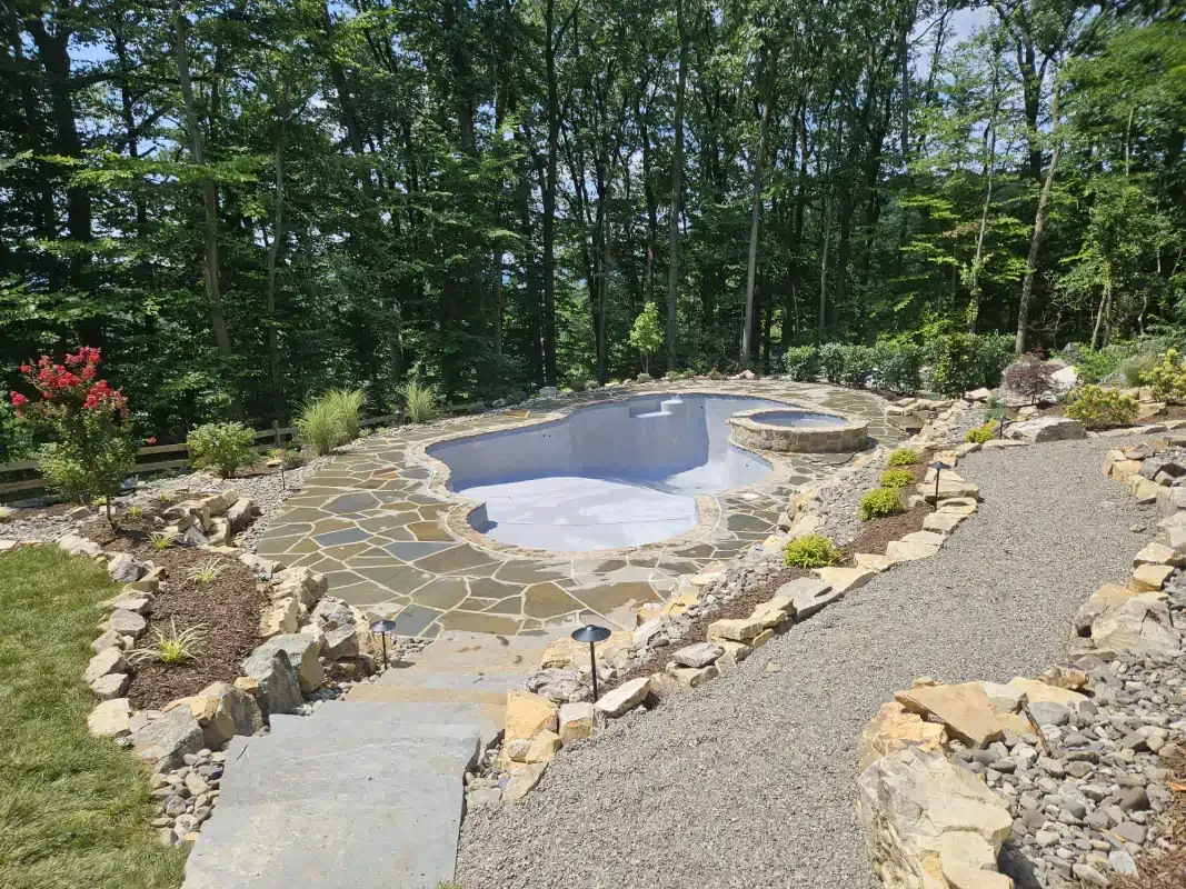 An empty swimming pool with a stone patio and gravel pathway, surrounded by trees and landscaping.