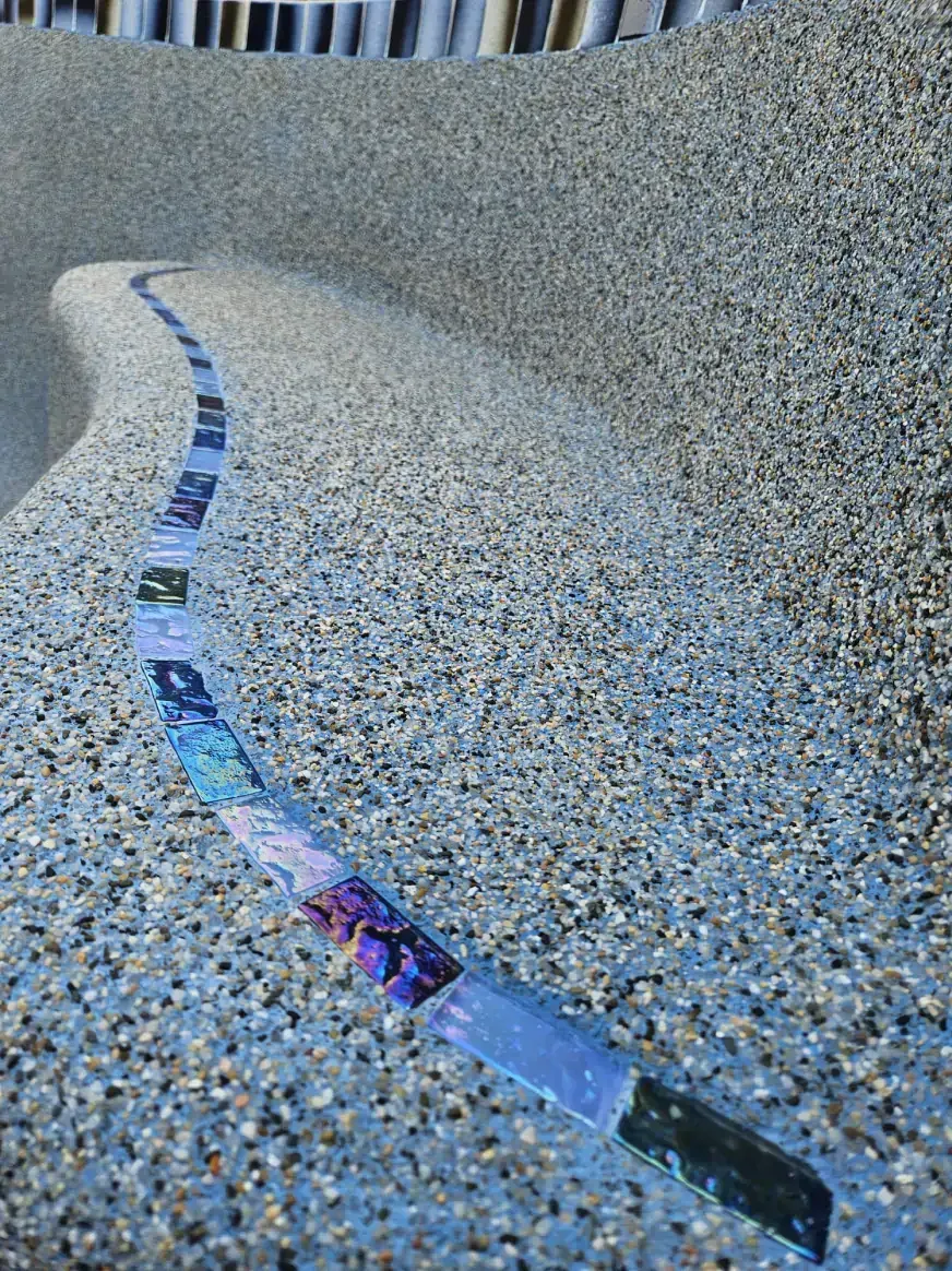 A curved, textured stone bench features a long, inlaid line of colorful, patterned mosaic tiles running along its surface.