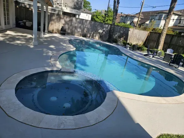 A curved backyard swimming pool with an attached circular hot tub, surrounded by a light-colored concrete patio and fence.